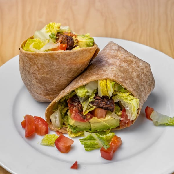 Two vegan wraps filled with lettuce, tomatoes, avocado, and plant-based meat on a white plate.
