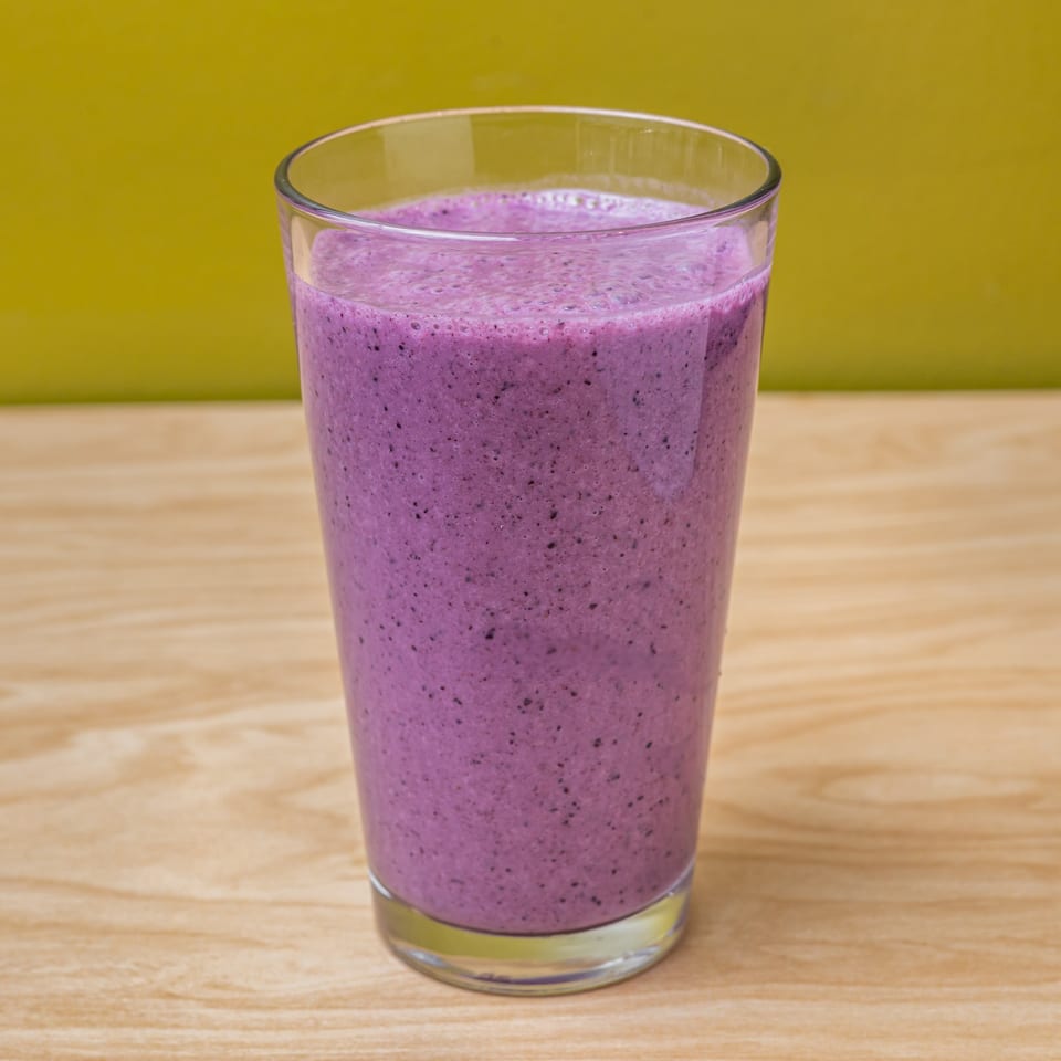A tall glass of vibrant purple vegan smoothie with small dark flecks sits on a wooden table.